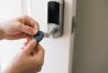 Hands holding a black key fob to a modern silver smart lock on a white door.